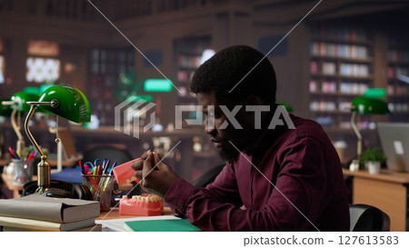 African american student examining fake teeth for dentistry thesis, working on stomatology research with appropriate tools in the campus library. Guy preparing for his career. Camera A. African american student examining fake teeth for dentistry thesis, working on stomatology research with appropriate tools in the campus library. Guy preparing for his career. Camera A. 127613583