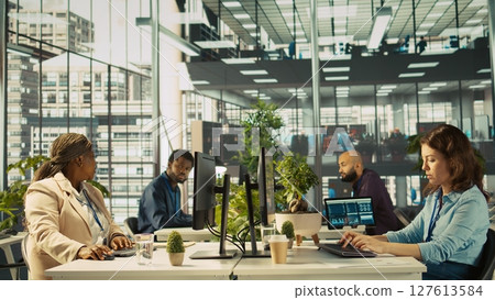 Accountant in busy modern startup agency imputing financial figures on PC after checking reports. Focused businesswoman working in modern office, crosschecking data, camera B 127613584