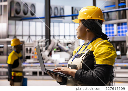 African american engineer in industrial warehouse uses laptop to monitor automated machinery. Industry 4.0 female expert controlling technological advancements in smart manufacturing facility. African american engineer in industrial warehouse uses laptop to monitor automated machinery. Industry 4.0 female expert controlling technological advancements in smart manufacturing facility. 127613609
