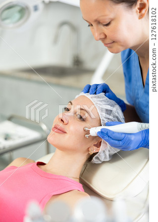 Experienced cosmetologist in rubber gloves does rf-lifting procedure on face for girl, smoothes wrinkles and tightens skin with use of electric hydrogen oxygen facial beauty machine in spa center 127618102