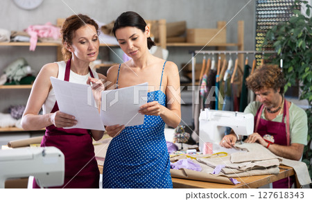 Two female dressmakers discussing dress sketches on paper. Man works on sewing machine. 127618343
