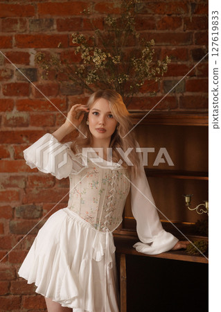Young woman in white dress standing barefoot near vintage cabinet against red brick wall Young woman in white dress standing barefoot near vintage cabinet against red brick wall 127619833