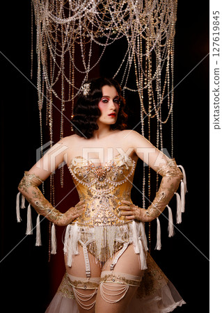 Cabaret dancer in vintage corset costume standing under chandelier on dark stage 127619845