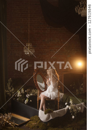 Blonde woman in white dress sitting by vintage mirror in moody room with brick wall background Blonde woman in white dress sitting by vintage mirror in moody room with brick wall background 127619846