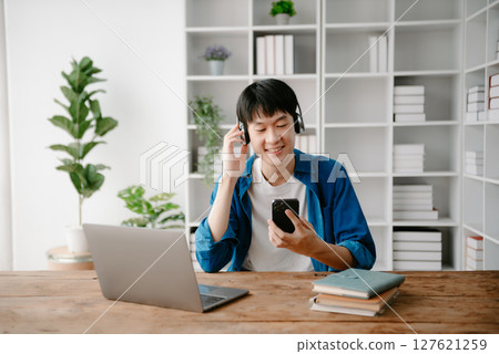 Smiling boy relaxing at home, she is playing music using smartphone tablet, laptop, and wearing white headphones. 127621259
