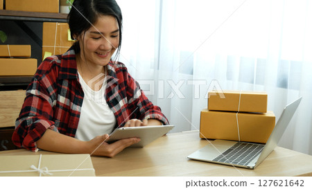 Asian woman startup small business at home office. Online seller entrepreneur young asian woman use tablet checking online order. Woman using tablet check stock goods supply delivery package shipping 127621642