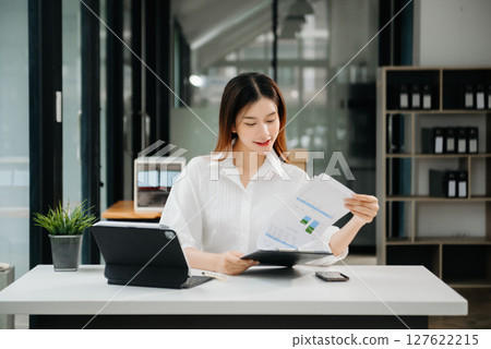 Young beautiful woman typing on tablet and laptop while sitting at the working white table modern office 127622215