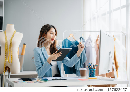 Fashion designer woman talking smart phone and using laptop with digital tablet computer in modern studio the clothes hanging on the racks Fashion designer woman talking smart phone and using laptop with digital tablet computer in modern studio the clothes hanging on the racks 127622457