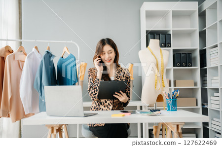 Asian young woman on desk in office of fashion designer and holds tablet, laptop and smartphone Asian young woman on desk in office of fashion designer and holds tablet, laptop and smartphone 127622469