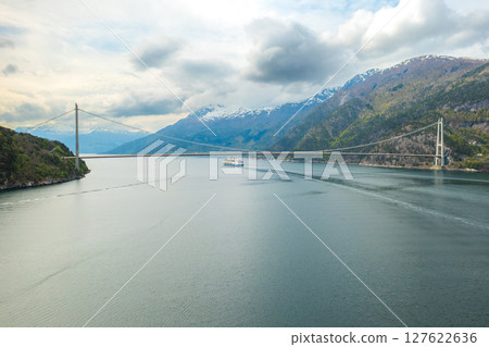 Hardanger Bridge gracefully arches over fjord calm waters with sailing sizable cruise liner. Majestic mountains encapsulates serene beauty of Norway Hardanger Bridge gracefully arches over fjord calm waters with sailing sizable cruise liner. Majestic mountains encapsulates serene beauty of Norway 127622636