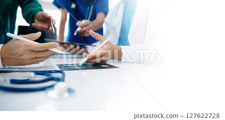 Medical team having a meeting with doctors in white lab coats and surgical scrubs seated at a table discussing a patients working online using computers 127622728
