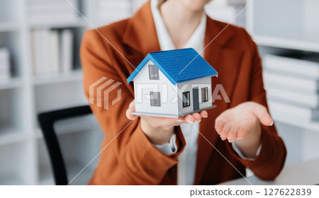 Young real estate agent worker working with laptop and tablet at table in office and small house beside it. Young real estate agent worker working with laptop and tablet at table in office and small house beside it. 127622839