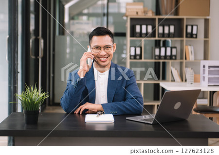 Young handsome man typing on tablet and laptop while sitting at the working wooden table modern office 127623128
