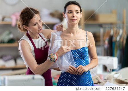 Woman seamstress trying on textile decoration on client dress in sewing workshop 127623247