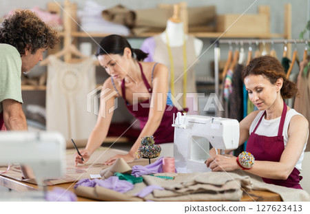 Female and male dressmakers drawing with curves and talking while other sewing Female and male dressmakers drawing with curves and talking while other sewing 127623413