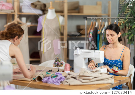 Woman seamstress sewing on a sewing machine. Female tailor drawing dress sketches on paper Woman seamstress sewing on a sewing machine. Female tailor drawing dress sketches on paper 127623414