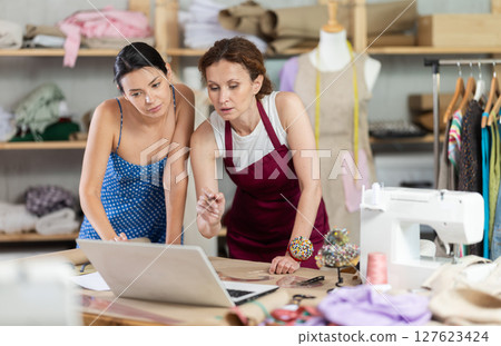 Woman tailor and female client discuss manufacture of clothes, look at laptop screen Woman tailor and female client discuss manufacture of clothes, look at laptop screen 127623424