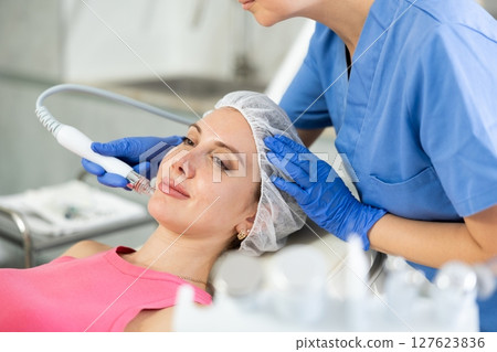 Young visitor receiving hardware electric massage and absorb blackheads and deeply clear skin by doctor in beauty clinic. Hardware cosmetology. Beautician hands in gloves makes face skin treatment for Young visitor receiving hardware electric massage and absorb blackheads and deeply clear skin by doctor in beauty clinic. Hardware cosmetology. Beautician hands in gloves makes face skin treatment for 127623836