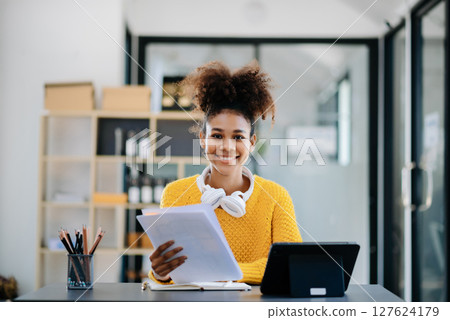 Young attractive Asian woman smiling thinking planning writing in notebook, tablet and laptop working from home, looking at camera at office 127624179