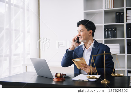 Asian male lawyer chatting online with client on laptop, smartphone  discussing details of lawsuit. in office 127624202