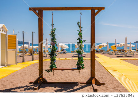 Decorative flower swings and empty beach loungers on a bright yellow path. Tranquil beach setup, summer rest and aesthetic vacation experience. 127625042
