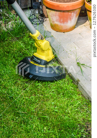 Cordless lawn trimmer cutting grass along patio edge near flower pots. High quality photo 127626380