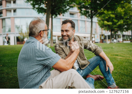 Father and adult son sitting on grass in park, sharing a story from the past. 127627156