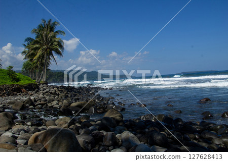 The charm of a rocky beach with clear blue waves and coconut tree vegetation on the shore in Sumatra, Lampung 127628413