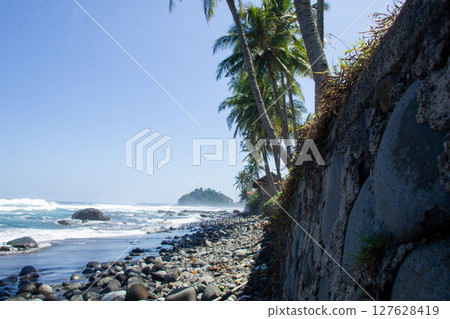Rocky Beach with Coconut Trees and Blue Waves Under Clear Sky, stone fence along the beach in Sumatra, Lampung 127628419
