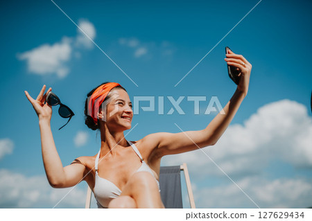 Young woman sitting by swimming pool outdoors in backyard garden, taking selfie. 127629434