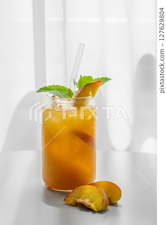 Peach drink or tea with ice, mint in a glass on a white table near window with fresh fruits  127629804