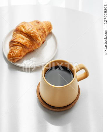 Coffee espresso in yellow cup and fresh croissant on white background with morning shadows. 127629808