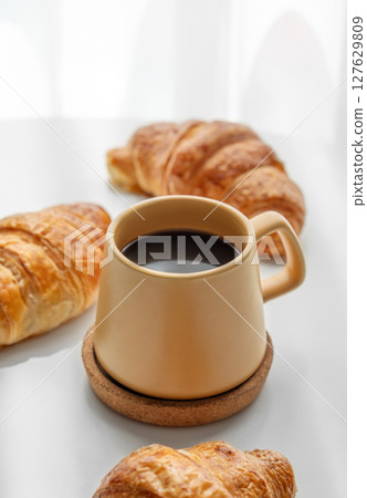 Coffee espresso in yellow cup and fresh croissants on white table near window with morning shadows. 127629809