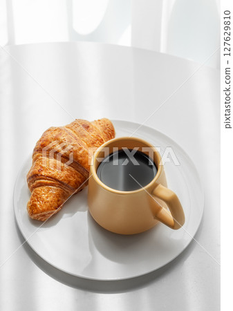 Coffee espresso in yellow cup and fresh croissant on a plate on white table near window 127629810