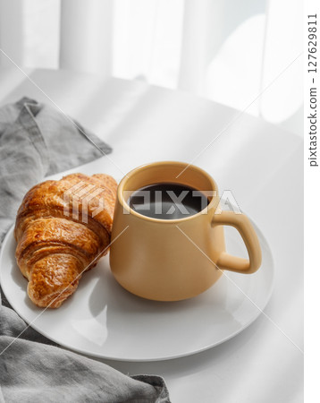 Coffee espresso in yellow cup and fresh croissant on a plate on white table near window Coffee espresso in yellow cup and fresh croissant on a plate on white table near window 127629811