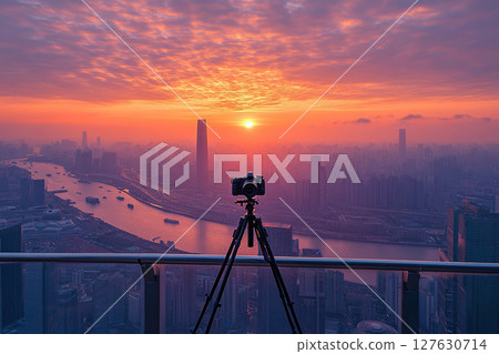 A tripod with a professional camera stands on a spacious balcony of a skyscraper at sunrise or sunset. 127630714