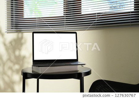 Laptop with blank screen on top of books on a black round table near window blinds Laptop with blank screen on top of books on a black round table near window blinds 127630767