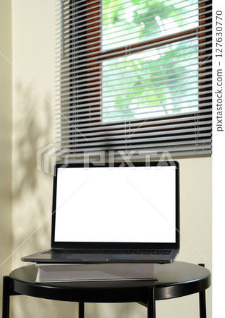 Laptop with blank screen on top of books on a black round table near window blinds 127630770