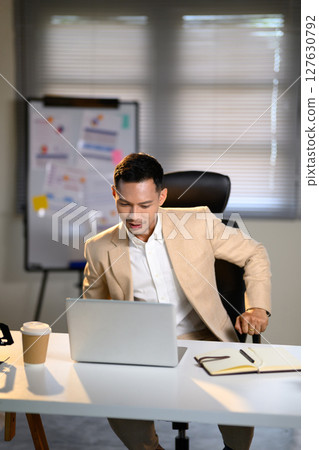 Confident businessman working on laptop at modern office desk with notebook and coffee Confident businessman working on laptop at modern office desk with notebook and coffee 127630792