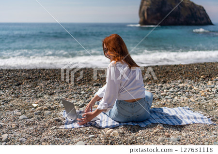 Beach laptop redhead: Woman uses laptop for work on pebble beach with ocean and cliff during daytime. Beach laptop redhead: Woman uses laptop for work on pebble beach with ocean and cliff during daytime. 127631118