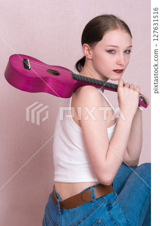 Young female musician confidently holds ukulele by neck and throws musical instrument over shoulder. She looks directly at camera. Beautiful Caucasian hipster woman has blank expression on her face 127631516