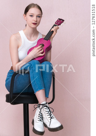 Young female musician strumming ukulele, sitting on bar stool and looking at camera while practicing musical instrument. Performer woman wearing white tank top, blue skinny jeans and white boots Young female musician strumming ukulele, sitting on bar stool and looking at camera while practicing musical instrument. Performer woman wearing white tank top, blue skinny jeans and white boots 127631518