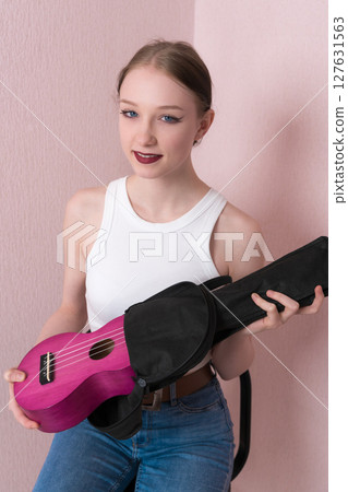 Young woman musician takes musical instrument ukulele out of its black soft case and looks into camera. Friendly 18 year old Caucasian female musician is wearing a white tank top and blue skinny jeans 127631563