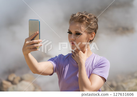 Beautiful female traveler using her cell phone to record self portrait to video call with friends in remote location against backdrop of steaming geysers in crater of an active volcano while traveling 127631606