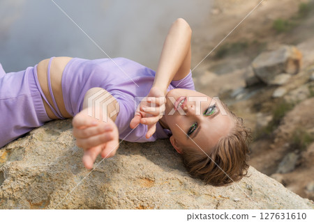 Friendly female fashion model lies on her back on large rock in crater of an active volcano, stretches her arms in front of her and looks at camera. Steam from geysers in crater volcano form backdrop 127631610