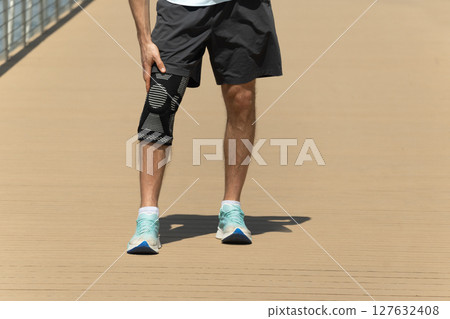 Man putting knee brace to support painful leg. Athlete bodybuilder leg wearing knee support brace or elastic kneecap bandage. Sleeve for compression. Outdoors. 127632408