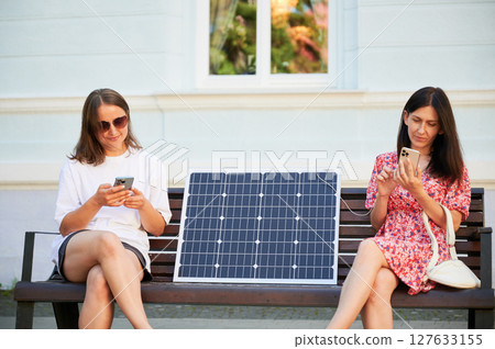 Two pretty women using portable solar battery to charge their mobile phones. Young female sitting on bench and charging their devices from solar panel. Green energy lifestyle for everyone. 127633155