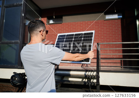 Back view of man holding solar panel toward the sun for charging electric mountain bike. Scene set against brick building. Back view of man holding solar panel toward the sun for charging electric mountain bike. Scene set against brick building. 127633156