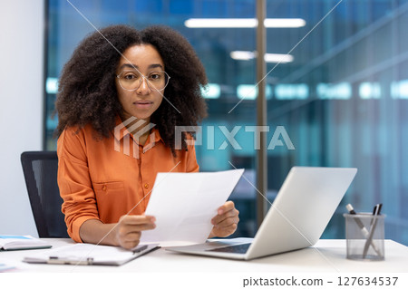 A serious woman with curly hair wears glasses and examines paperwork, with a laptop on a desk in an office. 127634537