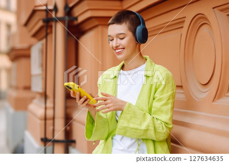 Woman wearing headphones posing outdoors, listens to music and walks along streets of a sunny city. Woman wearing headphones posing outdoors, listens to music and walks along streets of a sunny city. 127634635